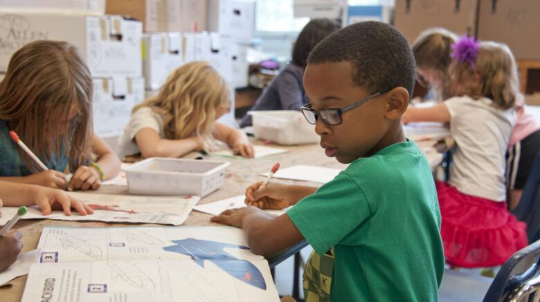Young children sit at desks working on an assignment for school. In the foreground, a boy wearing a green sweater and blue glasses looks at instructions to draw a picture.