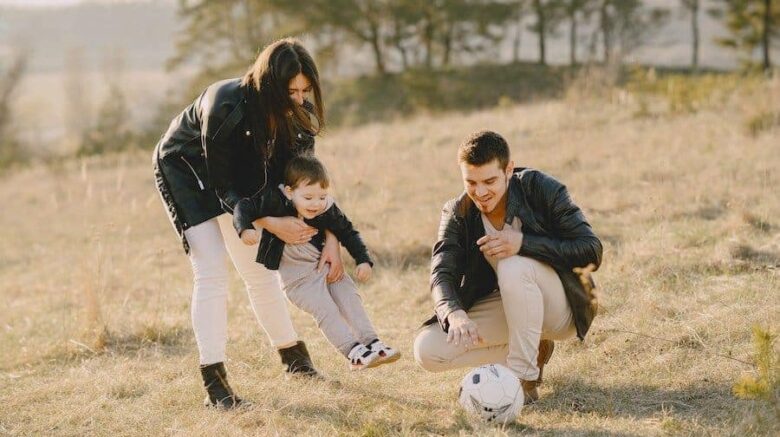 A man and woman play with a young boy in the field, the woman holding him as he looks at a soccer ball
