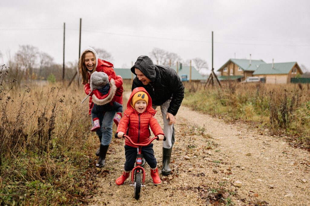 A young boy in a read winter jacket and boots and an orange hat rides on a bike while his parents walk behind him. His mom is carrying a younger child while his dad steadies the bike.