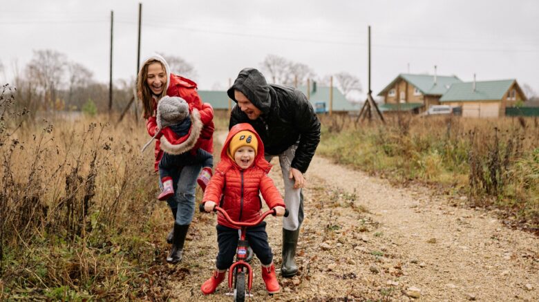A young boy in a read winter jacket and boots and an orange hat rides on a bike while his parents walk behind him. His mom is carrying a younger child while his dad steadies the bike.