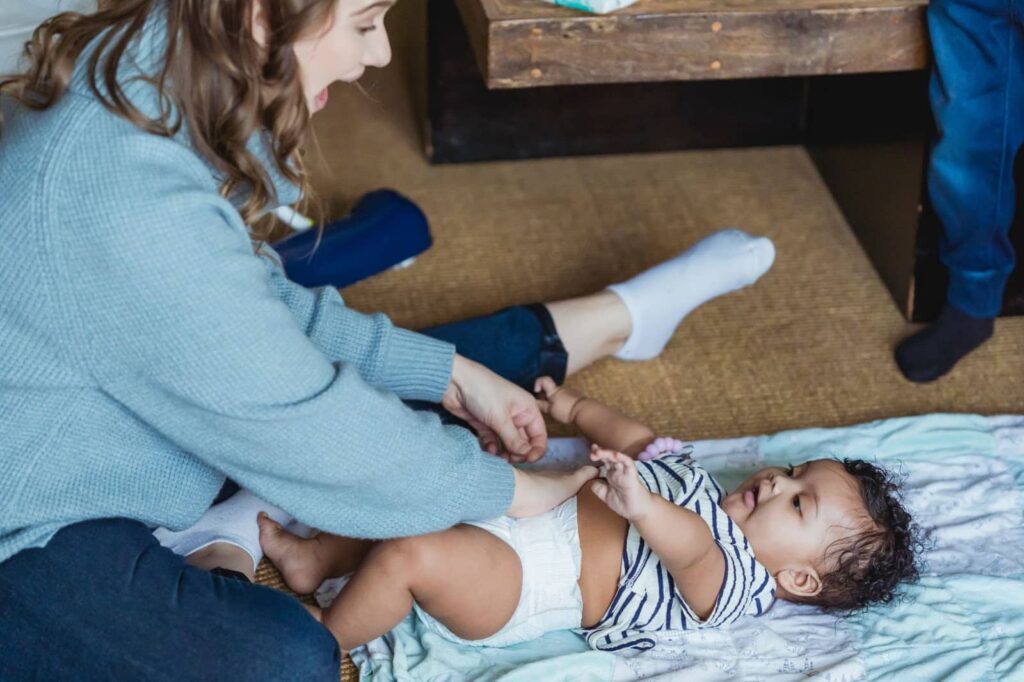 A woman sits facing a rug with a very young child laying on it, her hands reaching out towards the child as the two smile at each other.