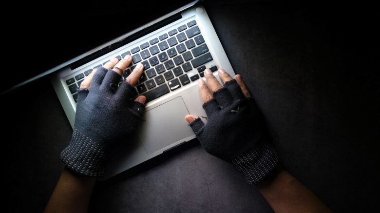 A pair of hands wearing dark gloves that don't cover the fingers rest against the keyboard of a computer, the light from the screen casting a dim glow.