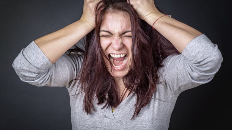 A woman wearing a grey shirt screams angrily, pulling on her hair in frustration