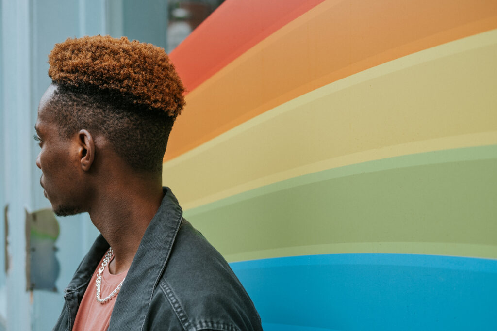 A man wearing a denim jacket over a pink shirt stands in front of a rainbow pattern