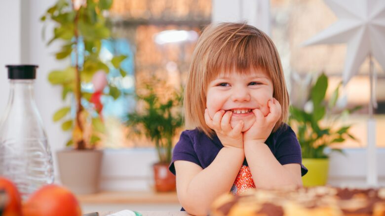 A young girl sits with her elbows resting on a table with a plate of food in front of her. Her hands are cupped around her cheeks and she is smiling while looking at something slightly to the left of the camera.