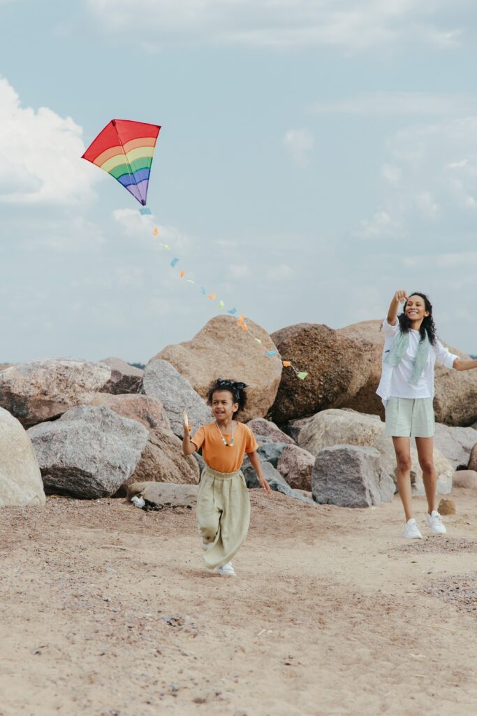 A young girl holds onto a rainbow kite while a woman watches in the background. The scene is set against some rocks.