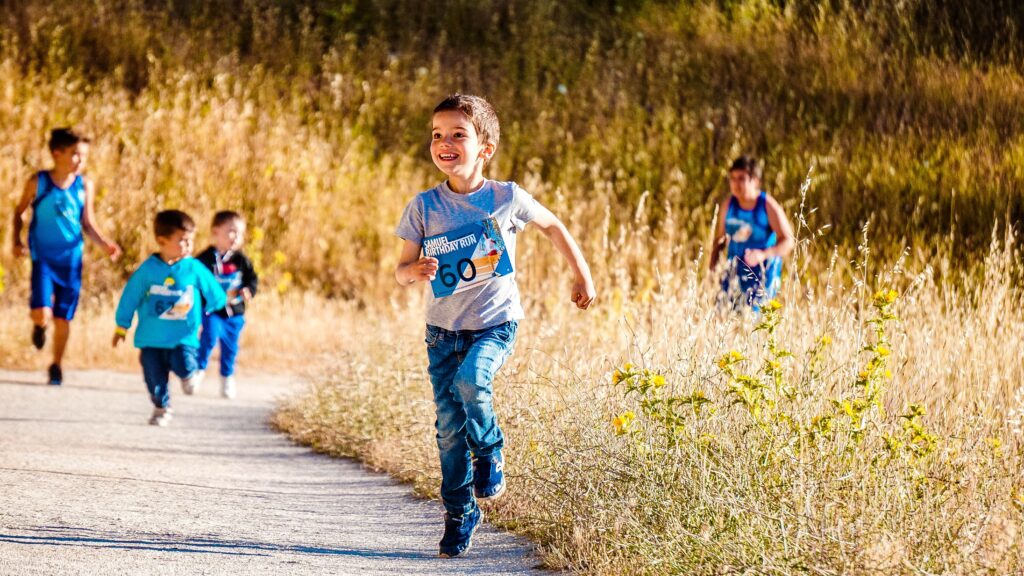 A group of boys is running in what appears to be a scaled-down marathon. In the foreground, a young boy in jeans and a t-shirt smiles while running along a path in between tall plants.