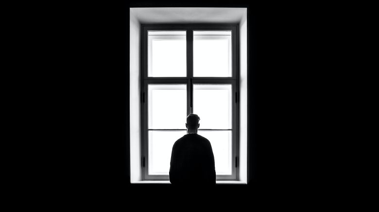 A man stands in shadow in front of a bright window, the walls around him shrouded in darkness.