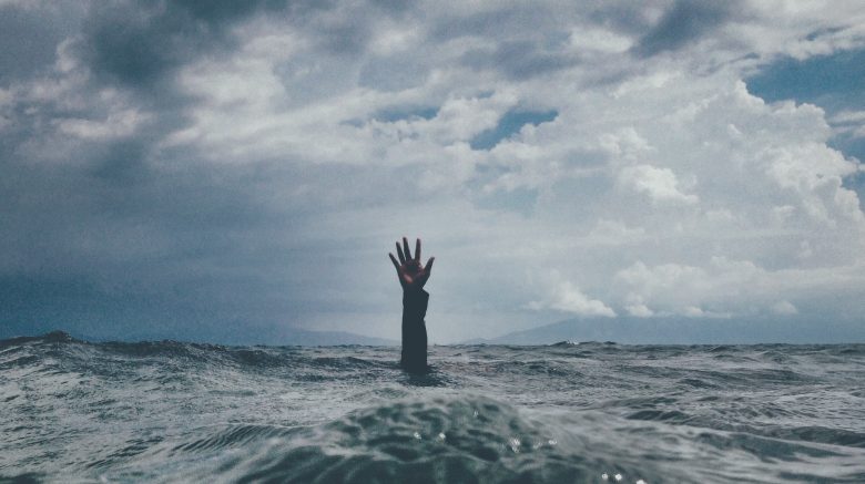 Beneath a cloudy sky, an outstretched hand emerges from ocean waves, the only visible sign of a person struggling to resurface as the current drags them down.
