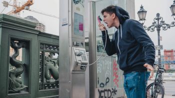 A man standing in front of a graffitied wall yells into an old payphone.