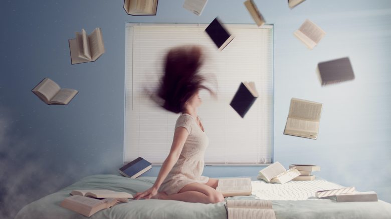 An action shot of a woman flipping her hair back while sitting on a bed. All around her, books are flying through the air, seemingly under their own power.