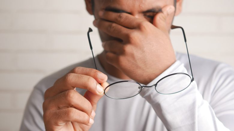 A man holds his glasses away from his face while rubbing his eyes.