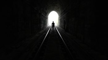A humanoid figure walks towards a bright light at the end of what appears to be a dark train tunnel.