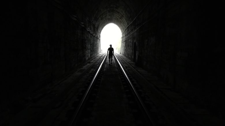A humanoid figure walks towards a bright light at the end of what appears to be a dark train tunnel.